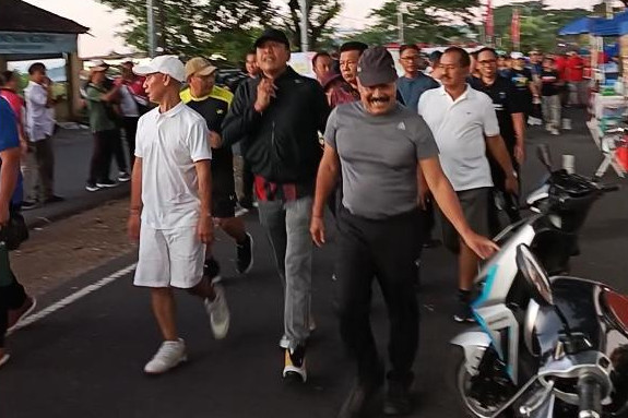 Sekelompok peserta, termasuk beberapa pejabat daerah, tampak bersemangat mengikuti kegiatan jalan santai di sepanjang jalan utama yang ditutup sementara. Suasana santai dan akrab terlihat dari senyum dan interaksi peserta, dengan latar belakang stand UMKM dan kendaraan listrik di sisi jalan.