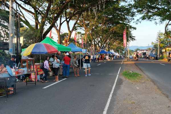 Deretan stan UMKM berjajar di pinggir jalan raya yang ditutup sementara untuk acara “Karangasem Akhir Pekan”. Beberapa warga tampak berbelanja dan berinteraksi di depan tenda berwarna-warni yang menjual makanan dan minuman. Pohon besar menaungi area sepanjang jalan, menciptakan suasana sejuk dan meriah.